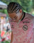 Person mit Cornrows, Brille, rosafarbenem T-Shirt mit "SPORADE" und "SBA" Logos, und einer Kreuzkette.