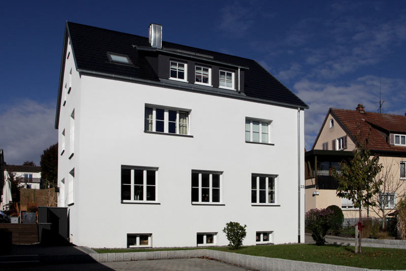 Altbau in der Wartenbergstraße: weiß verputztes Wohnhaus mit dunklem Satteldach, Gaube und Dachfenster, davor Rasen und gepflasterte Einfahrt