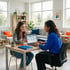 A woman in a blue shirt shows a laptop screen to a young girl sitting at a desk in a bright office or living room.