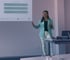 Frau mit hellblauem Blazer und weißer Hose präsentiert vor einer Projektionsfläche mit Diagramm.