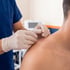 A practitioner in blue scrubs and white gloves performs dry needling or acupuncture on a patient's shoulder.