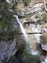 Wasserfall stürzt über graue, mehrschichtige Felsen in ein Becken, umgeben von Bäumen und Moos.
