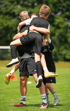 Drei junge Fußballspieler in schwarzen Trikots umarmen sich jubelnd auf einem grünen Rasen.