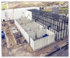 Baustelle einer großen Industrieanlage mit Kran, Fundamenten, Betonwänden und Stahlgerüsten unter bewölktem Himmel.