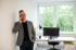 Ein Mann mit Brille und grauem Sakko telefoniert in einem Büro mit einem Schreibtisch, Computer und Fenster.