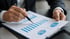 Hands holding a pen, pointing at blue bar graphs and pie charts on a white document on a clipboard.