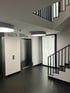 Flur mit Aufzug, schwarzem Fliesenboden, weißer Wand und Treppe mit schwarzem Geländer.