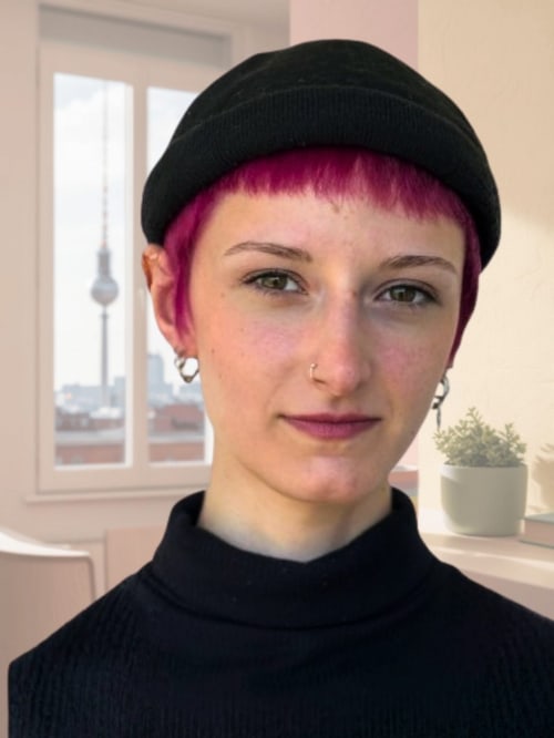Lächelnde Person mit kurzen pinkfarbenen Haaren, Mütze und Nasenring vor einem Fenster mit Blick auf den Berliner Fernsehturm.