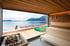 Moderne Holzsauna mit Panoramafenster, Blick auf einen See, Berge, eine Terrasse mit Liegestühlen und einen Saunaofen.