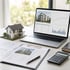 Ein Hausmodell, Laptop mit Immobilien-Charts, Taschenrechner und Baupläne liegen für eine Immobilienanalyse auf einem Tisch.