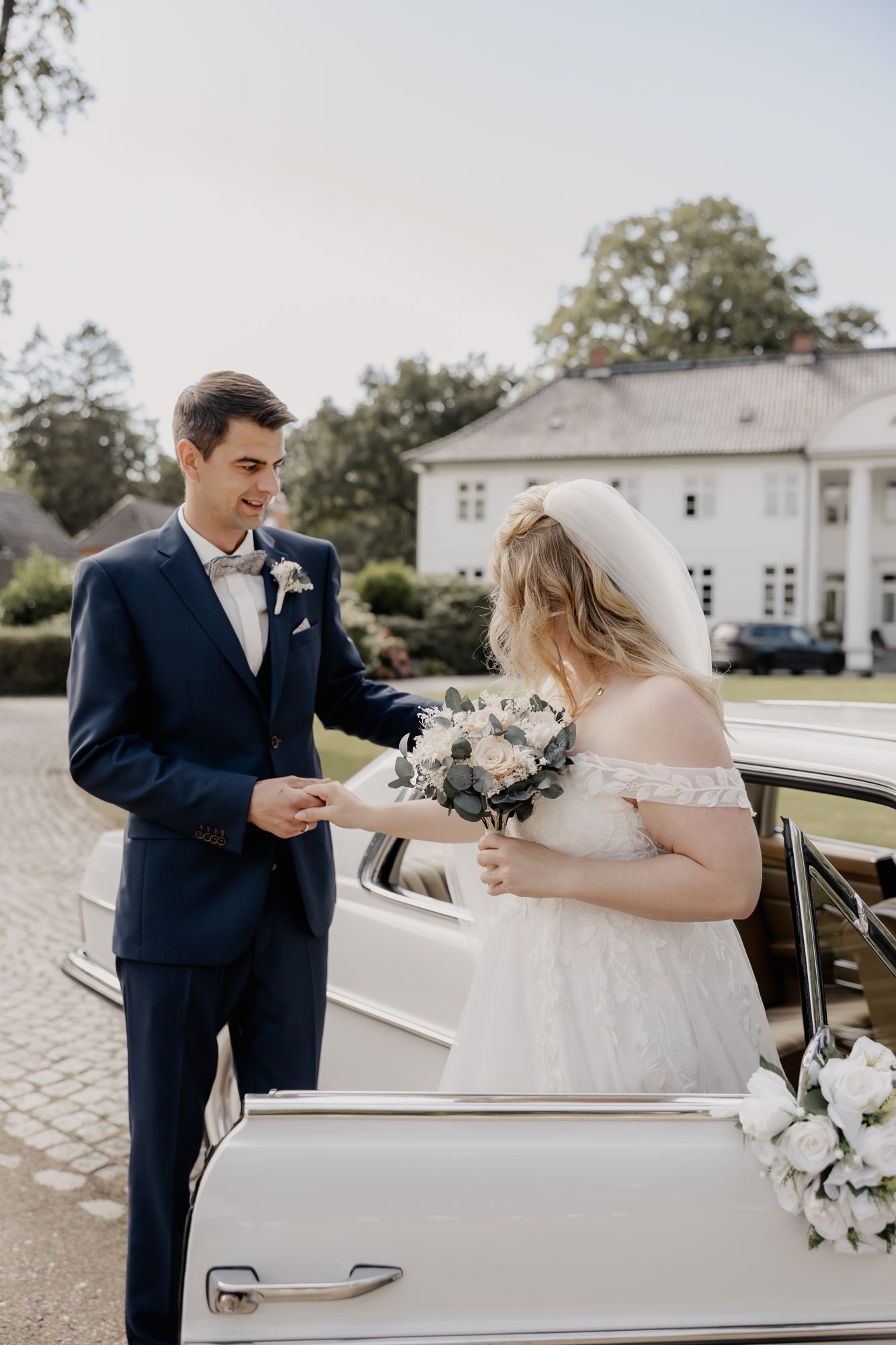 Bräutigam nimmt die Braut mit Brautstrauß an der Hand neben dem Hochzeitsauto