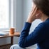 A woman in a blue sweater sits with her hand pressed against her forehead near a window with a mug and book.