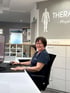 Eine lächelnde Mitarbeiterin mit Brille sitzt am Empfang einer Physiotherapie-Praxis vor einem Computerbildschirm.