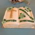 Konfirmationstorte in Buchform mit goldenem Kreuz, grünen Blumen und Schriftzug „VIVIEN 14.04.2024 ZUR KONFIRMATION“.