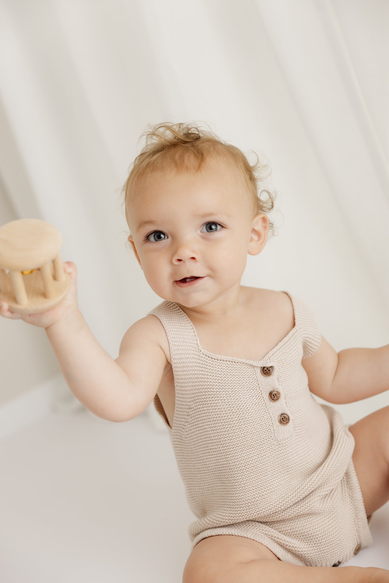 Baby sitzt vor weißem Vorhang und hält ein Holzspielzeug