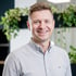 Lachender Mann mit kurzen Haaren und hellem Hemd vor verschwommenen Pflanzen.