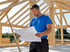 Zimmerer in blauem T-Shirt liest Baupläne auf einer Baustelle mit Holzrahmen.