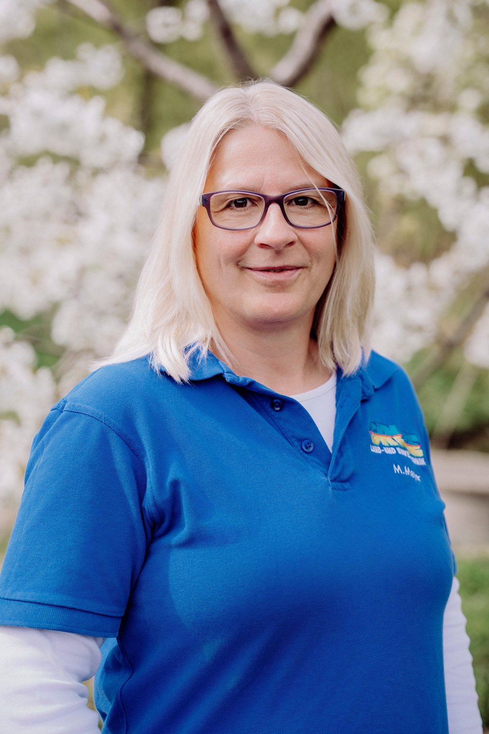 Blonde Frau mit Brille und blauem Poloshirt vor blühendem Baum lächelt.
