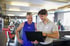 Ein Fitnesstrainer und ein Sportler betrachten gemeinsam einen Laptop im Fitnessstudio vor Trainingsgeräten.
