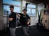 Zwei Männer in schwarzen T-Shirts und Mützen stehen in einem Fitnessstudio vor großen Fenstern.