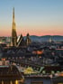 Wiener Skyline bei Sonnenuntergang mit Stephansdom, beleuchteten Gebäuden und Bergen am Horizont.