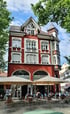 Rotes Gebäude mit weißer Fassade, Giebel, Turm und einem Café mit Außensitzplätzen und Sonnenschirmen.