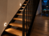 Moderne Holztreppe mit schwarzen Metallstreben und leuchtenden LED-Wandeinbaustrahlern zur Stufenbeleuchtung.