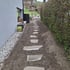 Ein neu angelegter Gartenweg mit flachen Trittsteinen in dunkler Erde zwischen einer Hauswand und einer hohen Hecke.
