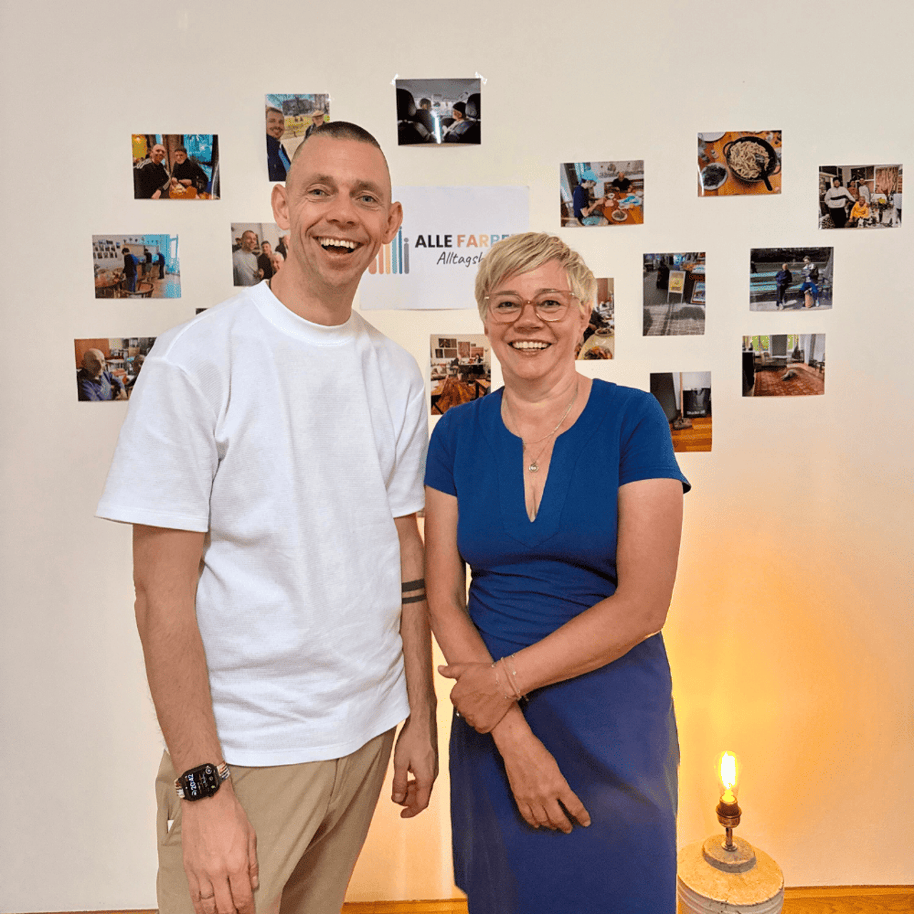 Mann und Frau lächeln vor einer Wand mit Fotos und einem "ALLE FARBEN" Poster.