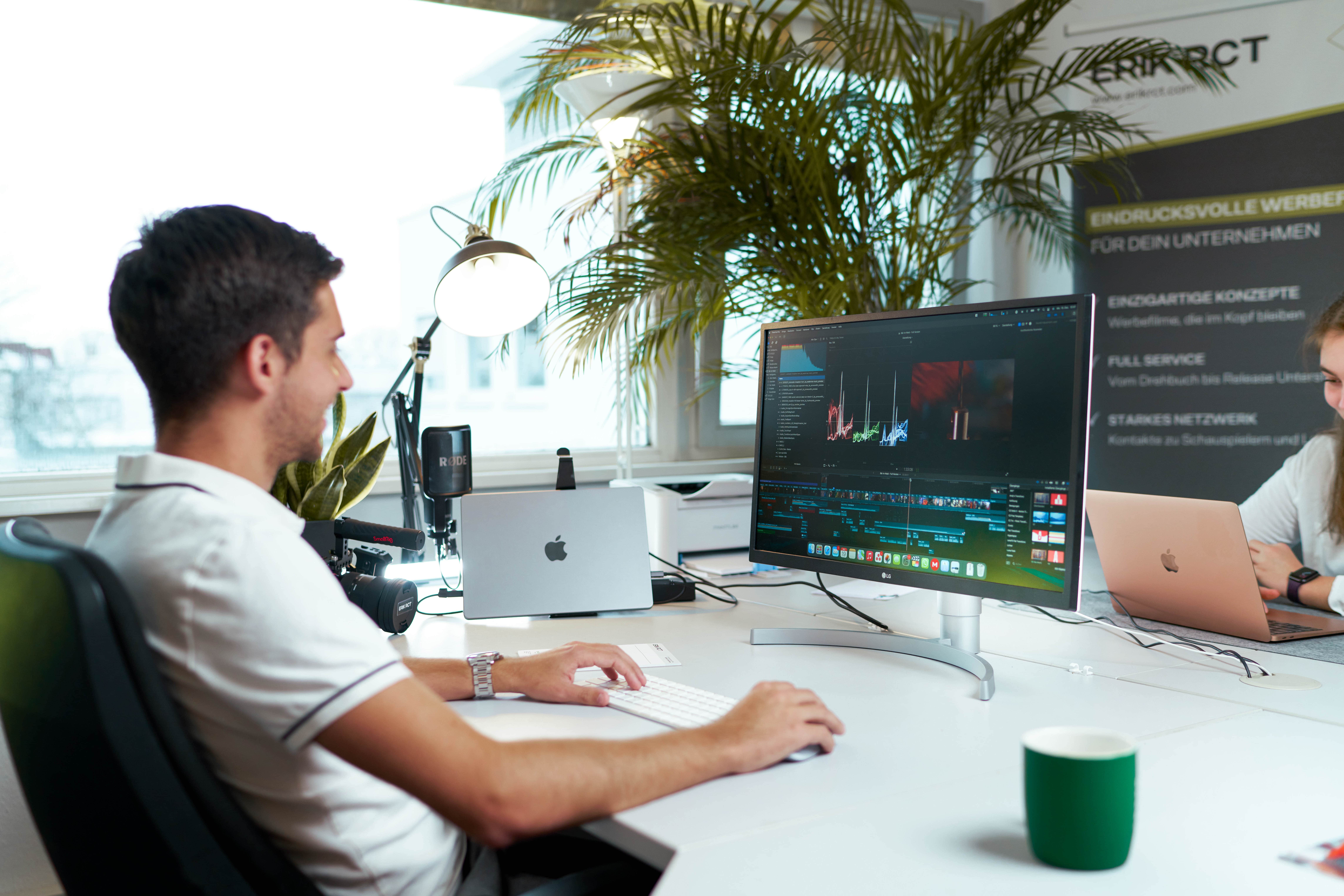 Ein Mann sitzt vor einem Computermonitor und bearbeitet Videos, während eine Frau am Laptop daneben sitzt.