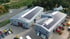 Zwei graue Gewerbegebäude mit Photovoltaik-Modulen auf den Dächern, umgeben von einem Parkplatz und Bäumen.