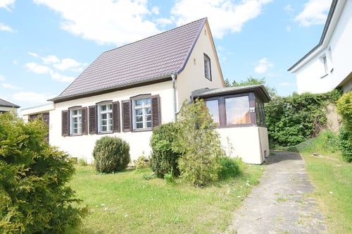 Beiges Einfamilienhaus mit braunem Dach, Fensterläden, Wintergarten, Garten und gepflasterter Einfahrt unter blauem Himmel.