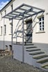 Gebäude mit grauer Tür, weißer Wand, grauen Stufen und einem Glasvordach, davor Kopfsteinpflaster.