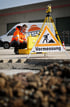 Ein Vermesser in orangefarbener Jacke beugt sich über eine gelbe Ausrüstungstasche, dahinter ein Vermessungsgerät und ein Warnschild "Vermessung".