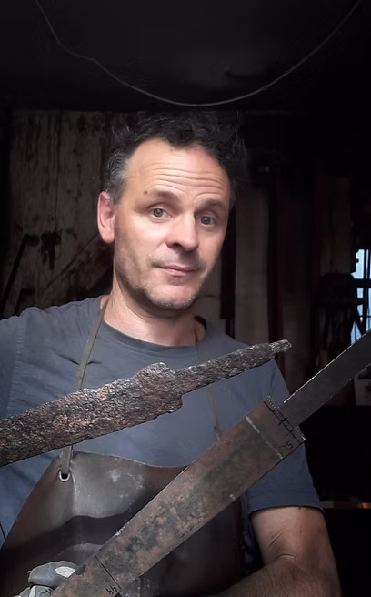 Ein Mann in Schürze hält ein rostiges, altes Schwert und eine neue, metallene Klinge mit Markierungen.