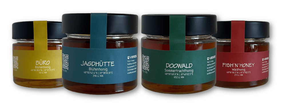 Vier Honiggläser mit schwarzen Deckeln und farbigen Etiketten: Gelb, Blau, Grün, Rot.