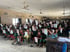 Zahlreiche Kinder in Uniform halten in einem Klassenzimmer weiße Geschenktüten hoch.