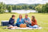 A family of four sits on a blanket for a picnic in a grassy field overlooking a lake and trees.