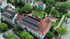 Luftaufnahme eines großen Gebäudes mit rotem Ziegeldach und Solarpaneelen, umgeben von Bäumen und Wohnhäusern.