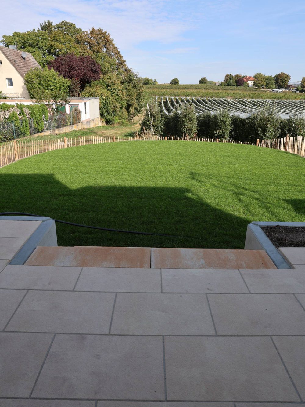 Blick von der Terrasse über die Treppe auf Rollrasen und Staketenzaun