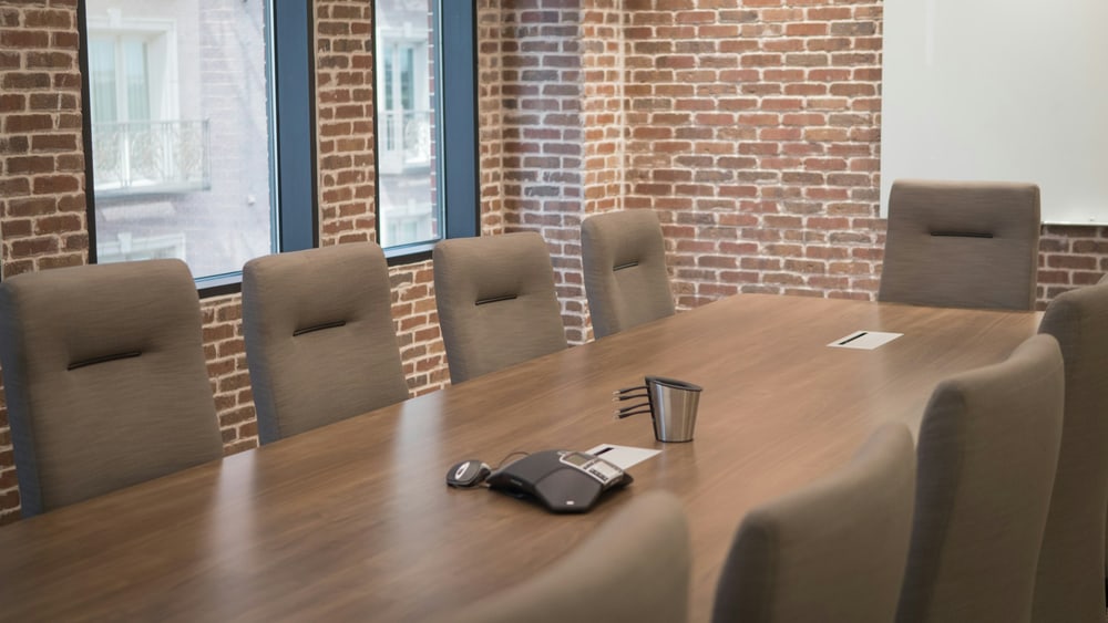Ein leerer Besprechungstisch mit gepolsterten Stühlen vor einer Backsteinwand, auf dem Tisch ein Becher und ein Konferenztelefon im Tageslicht.