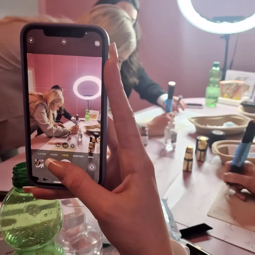 Eine Hand hält ein Smartphone, das ein Video einer Person aufnimmt, die an einem Tisch malt.