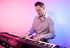 Ein Mann mit Brille spielt auf einem Keyboard unter blauen Bühnenlichtern, Bandmitglieder im Hintergrund.