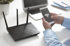 Person hält Smartphone mit WLAN-Symbol; schwarzer WLAN-Router und Laptop auf weißem Schreibtisch.
