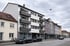 Modernes, weißes Wohngebäude mit grauen Balkonen und Geschäften im Erdgeschoss an einer Straße.
