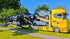 Gelber LKW-Autotransporter mit mehreren neuen Autos darauf, fährt auf einer Straße mit Bäumen und blauem Himmel.