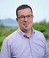 Ein lächelnder Mann mit Brille und Ziegenbart trägt ein lila Hemd vor einem unscharfen Hintergrund.
