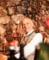 Lachender Mann mit Brille hält ein Glas Rotwein vor einer Steinwand mit Kerzen.
