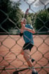 Tennisspieler in Aktion auf einem roten Sandplatz, gerade beim Aufschlag mit konzentriertem Blick und dynamischer Bewegung, umgeben von grünem Bewuchs und einem sonnigen Himmel.