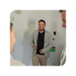 Ein lächelnder Mann in einem grauen Blazer mit einem Marker an einer weißen Tafel, umgeben von zwei Kollegen.